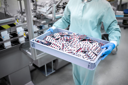 A young female worker in protective gear carefully carries a container filled with blister-packaged capsules, a crucial step in the transportation process of pharmaceutical products. Showcasing the importance of safety and attention to detail in the industry.