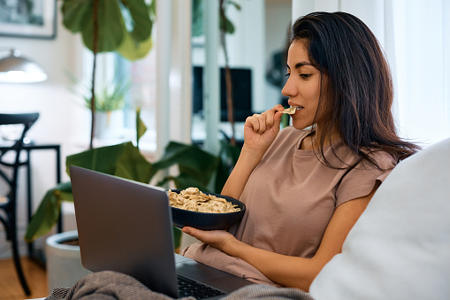Relaxed woman using laptop and eating snack while relaxing at home.