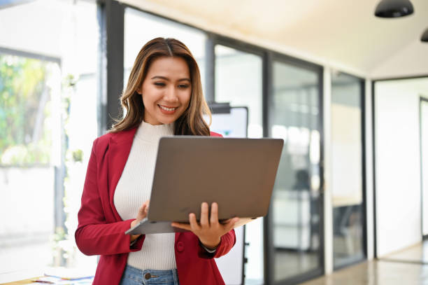 профессиональная азиатская бизнесвумен-миллениал стоит в своем офисе, используя портативный ноутбук - holding laptop women computer стоковые фото и изображения