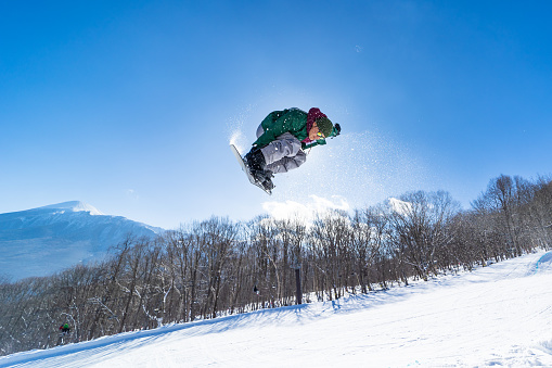 A Japanese male snowboard rider performing a jump on his snowboard on a clear sunny day back lit by the sun. Mt. Iwate can be seen in the background.