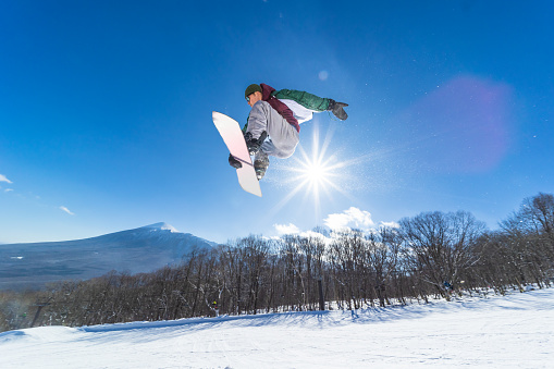 A Japanese male snowboard rider performing a jump on his snowboard on a clear sunny day back lit by the sun. Mt. Iwate can be seen in the background.