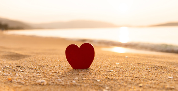 The figurine of the heart is in the sand on a luxurious sandy beach. Footprints in the sand. Waves in the sea. Dawn in the sky. Tropical layout for Valentine's Day.