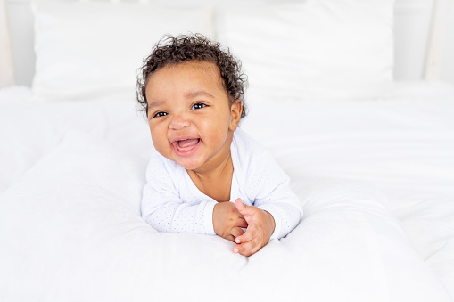smiling African-American little baby on white bed in bedroom laughing