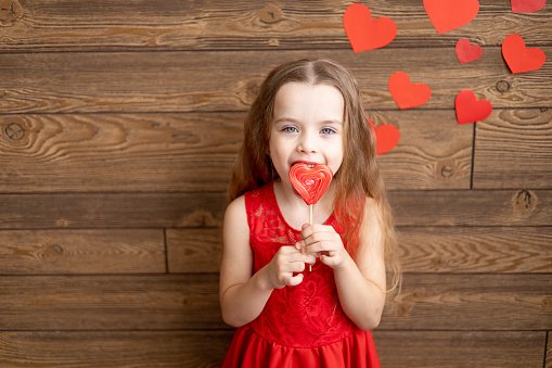 a little girl child in a red dress licks with her tongue a large lollipop in the form of a red heart on a dark brown wooden background with red hearts, the concept of Valentine's day, a place for text