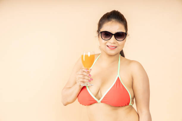 beautiful young brunette indian girl slim body wear red bikini and sunglasses enjoy glass of orange dink isolated on beige background, studio shot. tropical summer beach party and holiday concept. - party beach indian ethnicity adult imagens e fotografias de stock