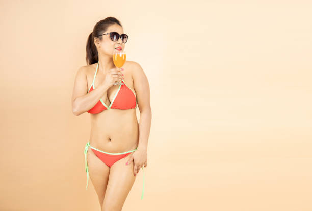 beautiful young brunette indian girl slim body wear red bikini and sunglasses enjoy glass of orange dink isolated on beige background, studio shot. tropical summer beach party and holiday concept. - party beach indian ethnicity adult imagens e fotografias de stock