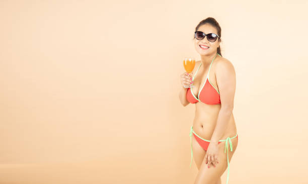 beautiful young brunette indian girl slim body wear red bikini and sunglasses enjoy glass of orange dink isolated on beige background, studio shot. tropical summer beach party and holiday concept. - party beach indian ethnicity adult imagens e fotografias de stock