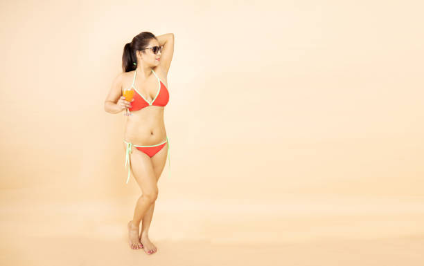 beautiful young brunette indian girl slim body wear red bikini and sunglasses hold glass of orange dink in hand isolated on beige background, studio shot. tropical summer beach holiday concept. - party beach indian ethnicity adult imagens e fotografias de stock