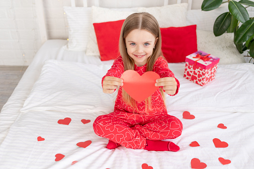 Valentine's day concept, a cute child girl is sitting on the bed at home in red pajamas and holding her heart in her hands and smiling, congratulating on the holiday