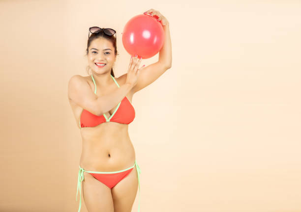 игривая молодая азиатская индианка стройное тело в красном бикини держит игру в пляжный мяч изолированно на бежевом фоне, студийный снимок - party beach indian ethnicity adult стоковые фото и изображения