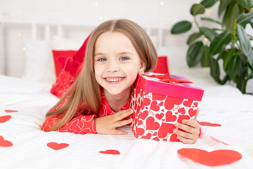 the concept of Valentine's day, a cute child girl is sitting on the bed at home in red pajamas and holding a gift in her hands and smiling or laughing with happiness, congratulating on the holiday