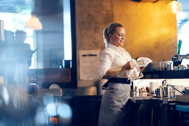A blond female professional bartender is cleaning glass in a bar while standing behind a bar counter. stock photo