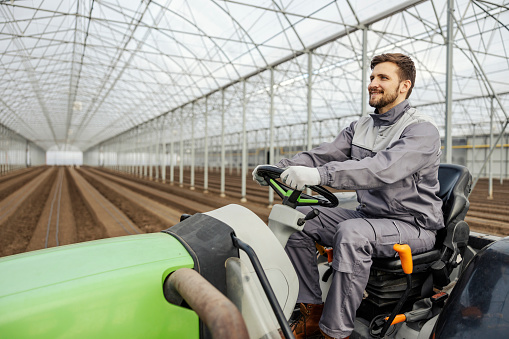 A smiling agronomy worker is cultivating the land and driving tractor in hothouse.
