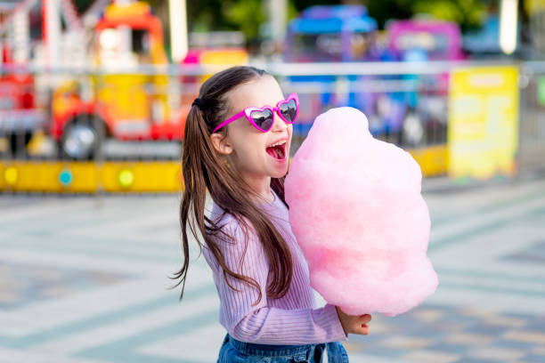 a child girl in an amusement park in the summer eats cotton candy near the carousels in sunglasses, the concept of summer holidays and school holidays a child girl in an amusement park in the summer eats cotton candy near the carousels in sunglasses, the concept of summer holidays and school holidays candyfloss stock pictures, royalty-free photos & images