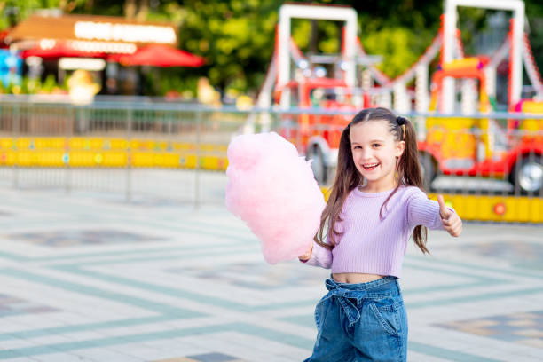 a child girl in an amusement park in the summer eats cotton candy near the carousels in sunglasses and shows the class, the concept of summer holidays and school holidays a child girl in an amusement park in the summer eats cotton candy near the carousels in sunglasses and shows the class, the concept of summer holidays and school holidays child cotton candy stock pictures, royalty-free photos & images