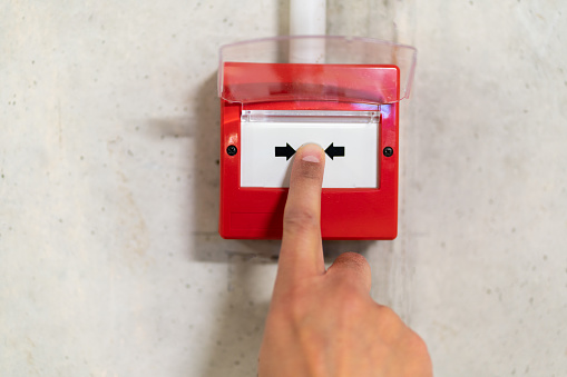 Close up of male hand pointing at red fire alarm switch on concrete wall in office building. Industrial fire warning system equipment for emergency.