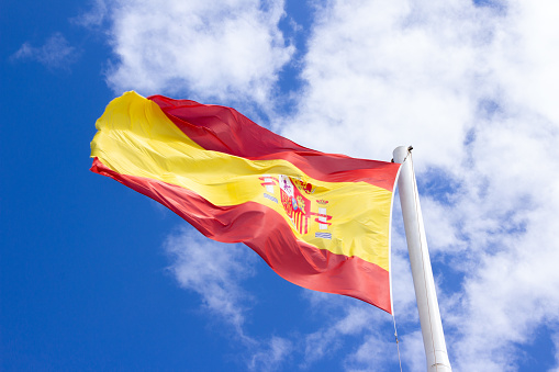 An isolated spanish flag in a really high flagpole. Rippling in the wind in a blue sky with some clouds.