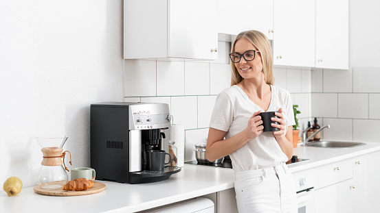 smiling female hold cup with espresso and looking on coffee machine standing at kitchen with light interior and modern household appliances