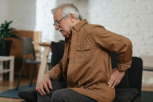 Worried senior man sitting on sofa at home and touching his back