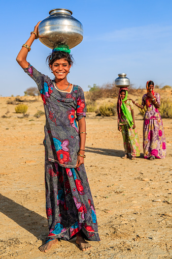 Indian young girls crossing sand dunes and carrying on their heads water from local well, Thar Desert, Rajasthan, India. Rajasthani women and children often walk long distances through the desert to bring back jugs of water that they carry on their heads.