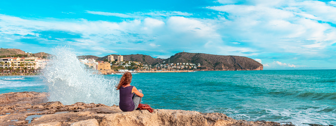 Woman sitting looking at waves crashing on rock ( Moraira,  Alicante province in Spain)