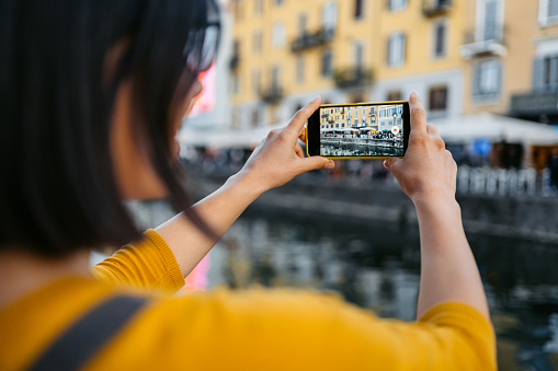 Beautiful young woman using her smart phone to take a photo of Navigli canal in Milan, Italy