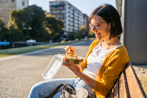 Beautiful young woman eating a bowl of salad while sitting on a park bench in Milan, Italy.