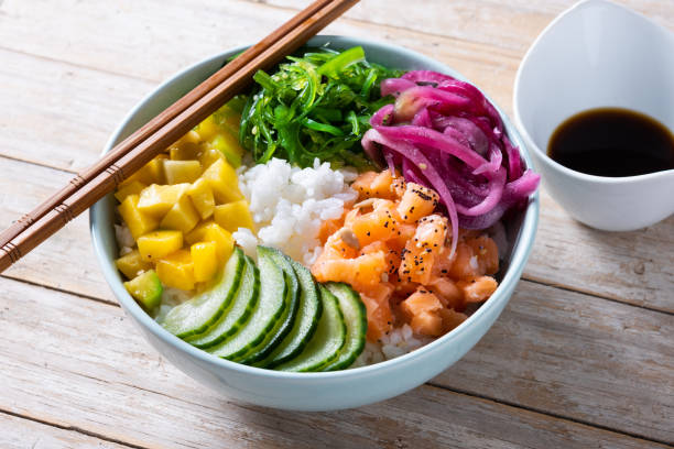 Poke bowl with rice, salmon,cucumber,mango,onion,wakame salad, poppy seeds ands sunflowers seeds on wooden background stock photo