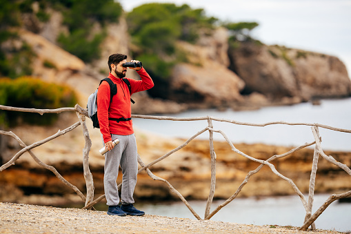 Male hiker with red shirt standing alone on a wooden fence in the landscape on the beautiful north coast of the island Mallorca looking into the distance to the sea with binoculars in his hands. Color editing. Part of a series.