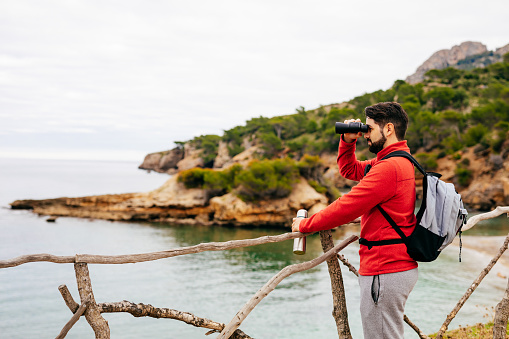 Hiker with red shirt standing alone on a wooden fence in the landscape on the beautiful north coast of the island Mallorca looking into the distance to the sea with binoculars in his hands. Color editing. Part of a series.