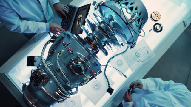 Top Down View of Two Engineers Discussing Work, Talking, Using Laptop Computer and Checking Blueprints in Jet Motor Manufacturing Facility. Scientists Researching Innovative Turbine Engine