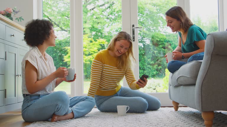 Three women relaxing in lounge at home drinking coffee and looking at photos or video on mobile phone - shot in slow motion