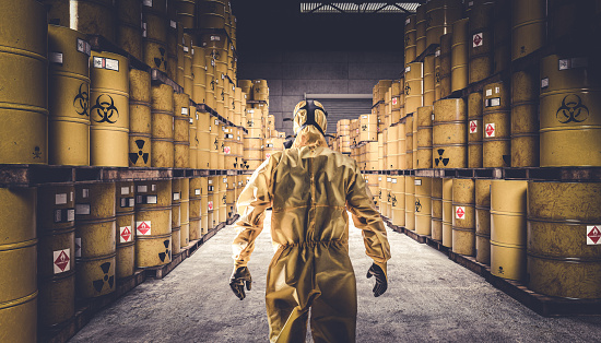 Man in yellow protective suit and gas mask, warehouse full of yellow metal barrels with danger symbols.