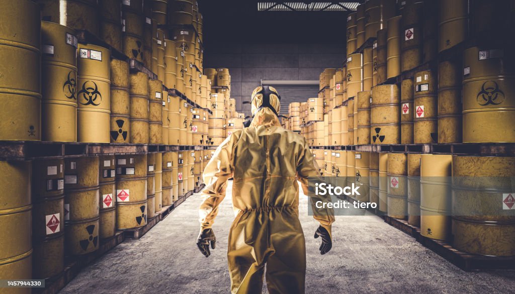 Man in yellow protective suit and gas mask, warehouse full of yellow metal barrels Man in yellow protective suit and gas mask, warehouse full of yellow metal barrels with danger symbols. Toxic Waste Stock Photo