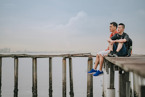 Asian Chinese down syndrome young man sitting side by side with father on wooden bridge seaside during overcast morning