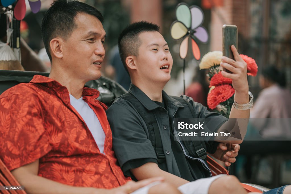 Asian Chinese down syndrome young man enjoying photographing with smart phone rickshaw ride in Penang street with father Active Lifestyle Stock Photo
