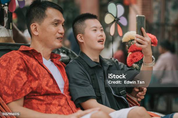 Asian Chinese Down Syndrome Young Man Enjoying Photographing With Smart Phone Rickshaw Ride In Penang Street With Father Stock Photo - Download Image Now