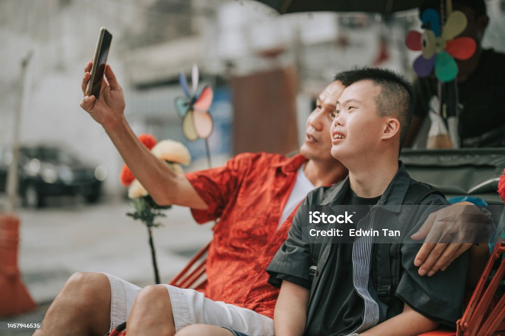 Asian Chinese down syndrome young man enjoying selfie  rickshaw ride in Penang street with father Asian Chinese down syndrome young man enjoying rickshaw ride in Penang street with father Active Lifestyle Stock Photo