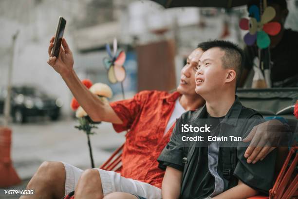 Asian Chinese Down Syndrome Young Man Enjoying Selfie Rickshaw Ride In Penang Street With Father Stock Photo - Download Image Now