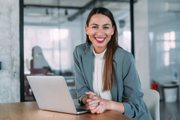 Successful businesswoman in modern office working on laptop. Portrait of elegant businesswoman sitting in her office looking at camera while using laptop. sales executive stock pictures, royalty-free photos & images