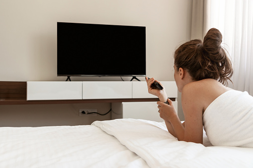 Brunette woman wrapped in towel lying on bed in bed room and watching television using tv remote. Blank screen tv.