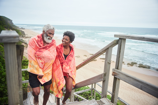 Smiling mature couple in swimsuits and wrapped in towels walking up steps by the ocean after a swim