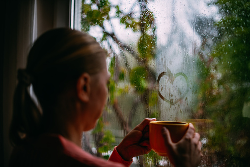 A Woman Drinking Coffee and Drawing Heart on the Window in a Rainy Day