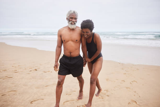 istock Mature couple in swimwear laughing while walking on a sandy beach 1457954901