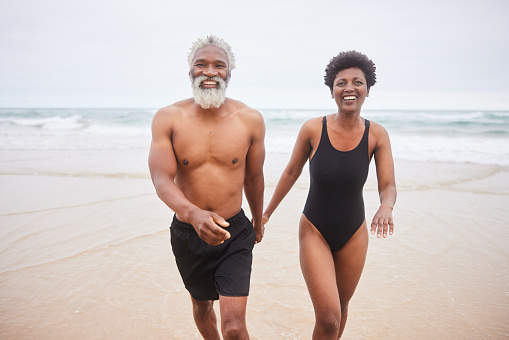 Smiling mature couple in swimwear walking hand in hand on a beach on an over cast day