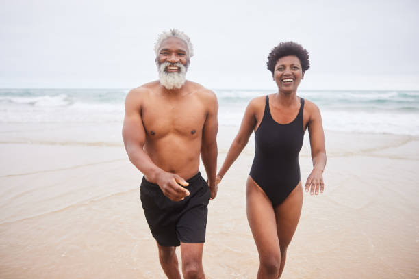 Mature couple in swimsuits smiling while walking on a sandy beach Smiling mature couple in swimwear walking hand in hand on a beach on an over cast day black woman bathing suit stock pictures, royalty-free photos & images