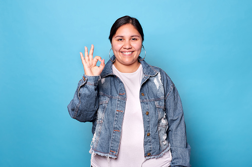 Young curvy latina woman wearing denim jacket and hoop earrings, smiling showing okay sign looking at camera isolated on blue background. Copy space.