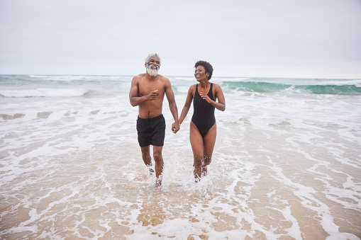 Laughing mature couple in swimwear walking hand in hand in the ocean surf on an over cast day