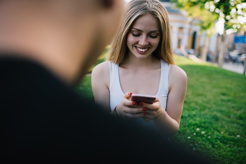 Happy youthful blond female chatting on smartphone and enjoying pastime with friend while sitting on grass during meeting in park