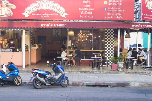 Thai people are eating outside of restaurant located at Wanghin Road in Bangkok ladprao. In center a couple is sitting. At left side staff is working. At right side out of restaurant a group of young people is sitting. On street motorcycles are parked